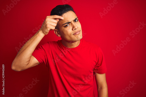 Young brazilian man wearing t-shirt standing over isolated red background pointing unhappy to pimple on forehead, ugly infection of blackhead. Acne and skin problem