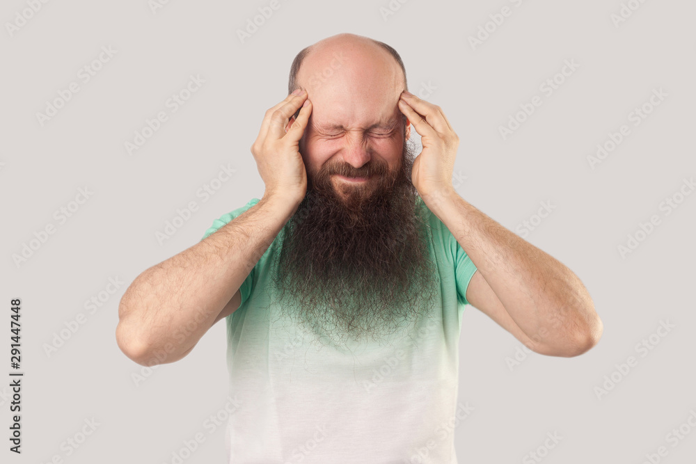 Naklejka premium Headache, thinking or confusion. Portrait of middle aged bald man with long beard in light green t-shirt standing and holding his painful head. indoor studio shot, isolated on grey background.