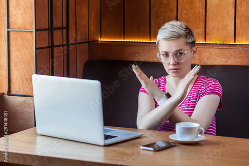 Portrait of warning aggressive young girl with blonde short hair in pink t-shirt is sitting in cafe and working on laptop and showing crossing raised arms like stop gesture. Indoor, looking at camera photo