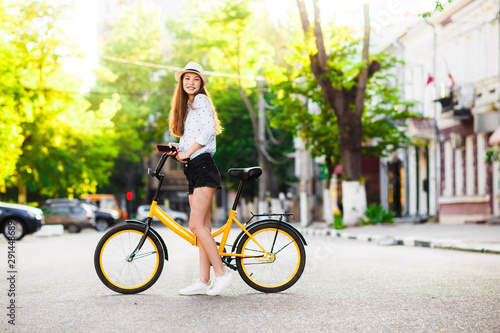 Girl teenager in a hat with a yellow bike walks through the summer city. © galina_kovalenko