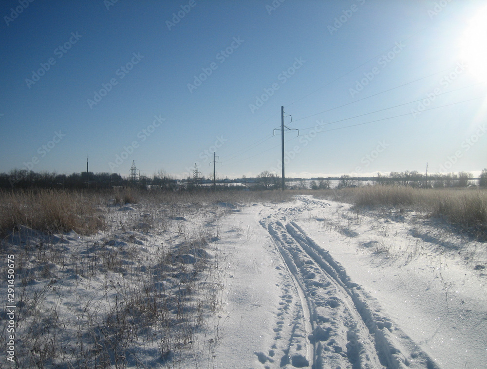 Winter road in a clean flat field on a clear frosty day. The sky is clear, blue and transparent. Snow covered the field in an endless carpet. The ruts of the road cut through the snowy surface.