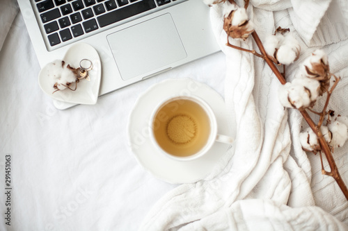 White mug with tea, cotton, laptop, plaid on the bed. Breakfast in bed. Cozy. Autumn. Winter.