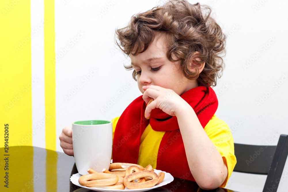 Little curly boy drinking from cup of hot drink tea in cold weather, sitting at home in scarf in kitchen at the table Little curly boy drinking from cup of hot drink tea in cold weather, sitting at home in scarf in kitchen at the table