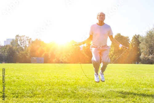 Active senior man doing jumping rope work out in park with yellow lens flare in background