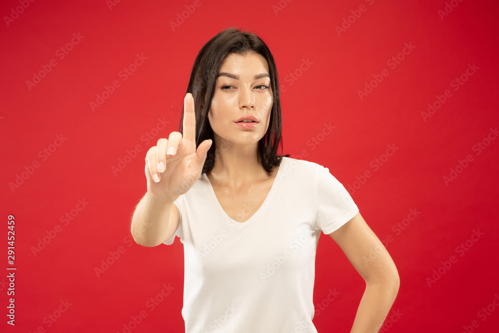 Fototapeta premium Caucasian young woman's half-length portrait on red studio background. Beautiful female model in white shirt. Concept of human emotions, facial expression. Touching empty search bar, copyspace.
