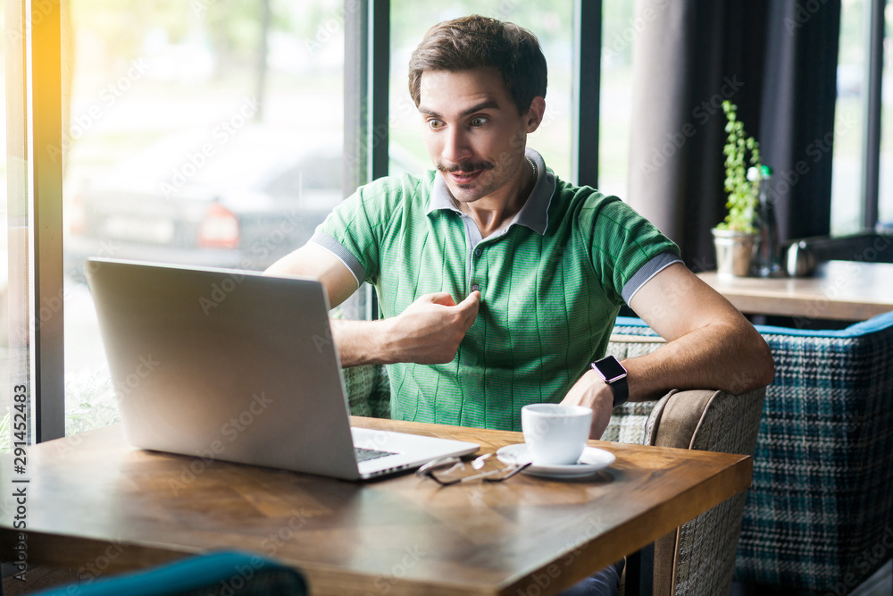 Who? me? Young amazed businessman in green t-shirt sitting, pointing and looking at laptop display with surprised face, asking. business and freelancing concept. indoor shot near big window at daytime Who? me? Young amazed businessman in green t-shirt sitting, pointing and looking at laptop display with surprised face, asking. business and freelancing concept. indoor shot near big window at daytime