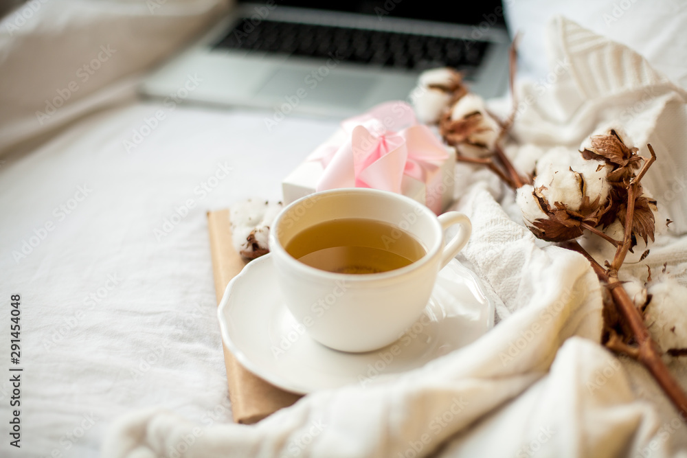 White mug with tea, cotton, notebook, plaid, book and white gift box with a pink ribbon on the bed. Breakfast in bed. Cozy. Autumn. Winter.
