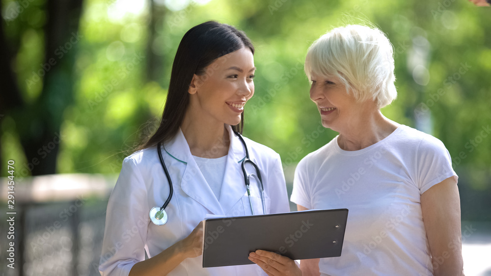Smiling nurse showing elderly woman examination results in hospital park Smiling nurse showing elderly woman examination results in hospital park