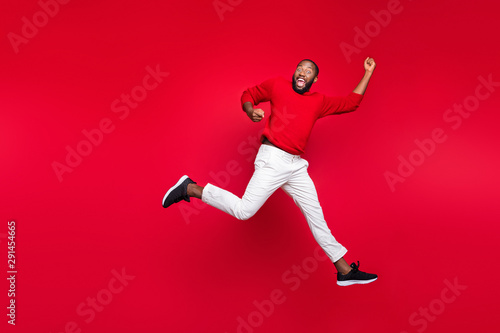 Full length photo of cheerful guy jumping raising hands screaming wearing pullover isolated over red background
