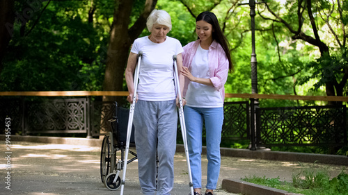 Slika na platnu Female volunteer helping disabled senior woman walk with frame in park, support