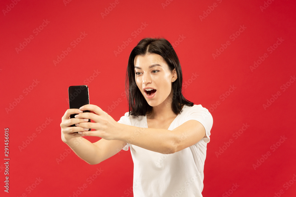 Caucasian young woman's half-length portrait on red studio background. Beautiful female model in white shirt. Concept of human emotions, facial expression. Making selfie or her own vlog, blog. Caucasian young woman's half-length portrait on red studio background. Beautiful female model in white shirt. Concept of human emotions, facial expression. Making selfie or her own vlog, blog.