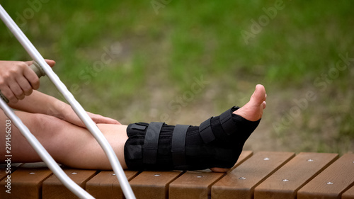 Slika na platnu Female with crutch sitting bench with ankle brace on leg, bone fracture, strain