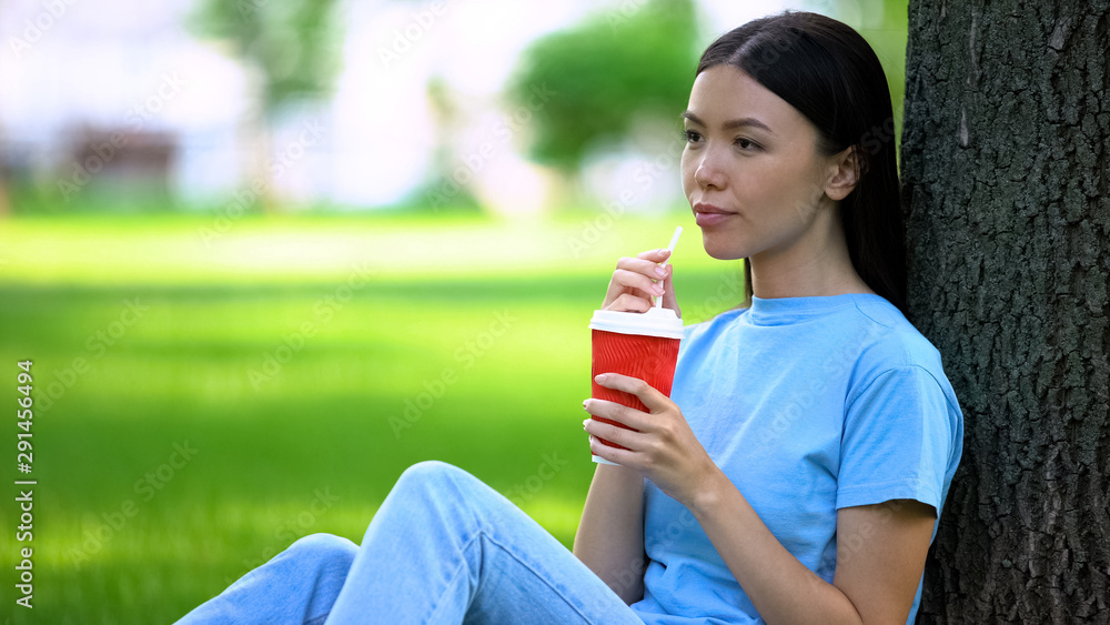 Happy female drinking lemonade through straw sitting garden tree, refreshment Happy female drinking lemonade through straw sitting garden tree, refreshment
