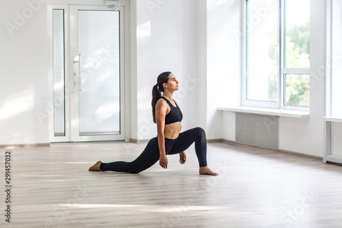 Young beautiful sporty woman wearing black sportswear practicing sport exercises, doing lunges near window in gym, stretching legs and standing on ashtanga pose, Indoor, sports lifestyle