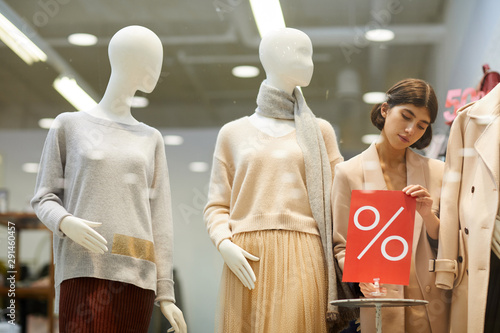 Fototapeta Portrait of beautiful saleswoman fixing red SALE sign and mannequins dressed Aut
