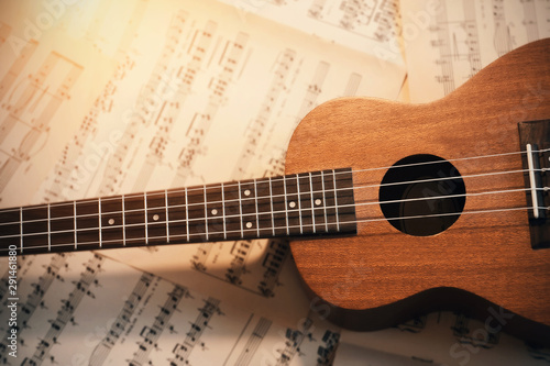 A small wooden ukulele with nylon strings lies on scattered sheets of musical notes, illuminated by the light 