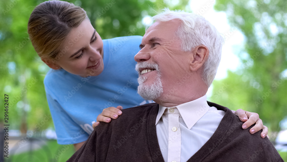 Smiling female volunteer comforting handicapped elderly man at hospital park Smiling female volunteer comforting handicapped elderly man at hospital park