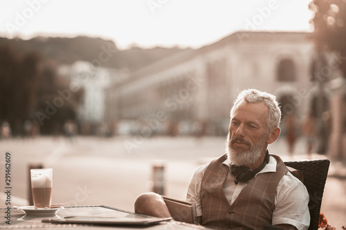 Stylish freelance businessman in casual clothes portrait. Handsome bearded man make a note in notebook or laptop. Male model drink coffee in cafe on city street