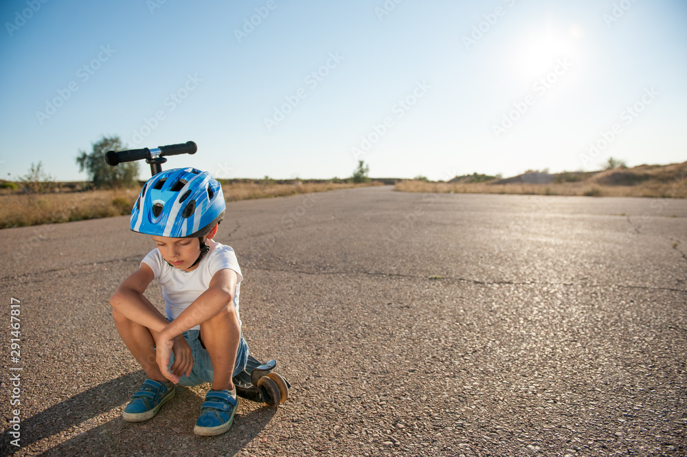 sad little caucasian boy in helmet sitting on his scooter on asphalt empty  road during summer hot sunset in California with copy space Stock Photo |  Adobe Stock