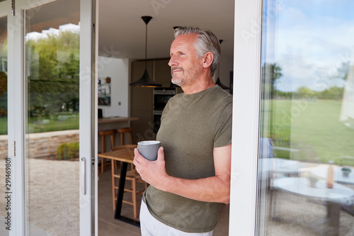 Senior Man Standing And Looking Out Of Kitchen Door Drinking Coffee photo