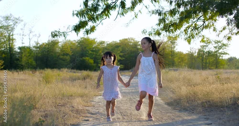 Two pretty beautiful little girls four and seven years old in white summer dresses, skipping along a dusty country trail holding hands on warm summer evening. Slow motion 4k 50 fps Two pretty beautiful little girls four and seven years old in white summer dresses, skipping along a dusty country trail holding hands on warm summer evening. Slow motion 4k 50 fps