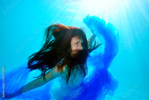 Surreal underwater portrait of the bride with her hair down, who floats near the bottom of the pool with a blue cloth in her hands in a white wedding dress. Unusual wedding. Fashion portrait.