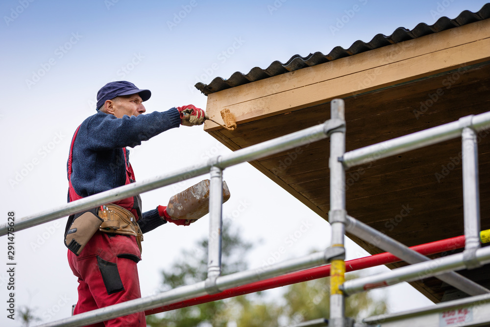 man on scaffolding painting house roof planks with paint roller
