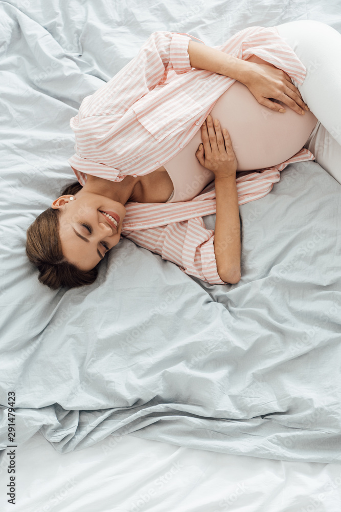 overhead view of smiling pregnant woman lying in bed and touching belly overhead view of smiling pregnant woman lying in bed and touching belly