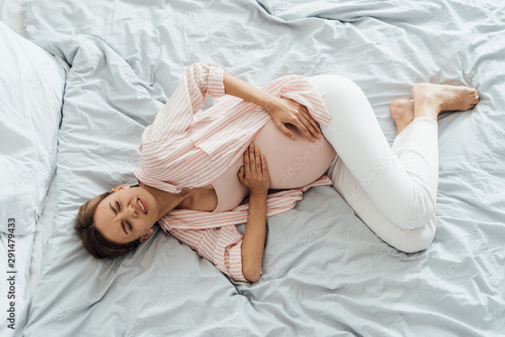 overhead view of pregnant woman lying in bed with grimace and touching