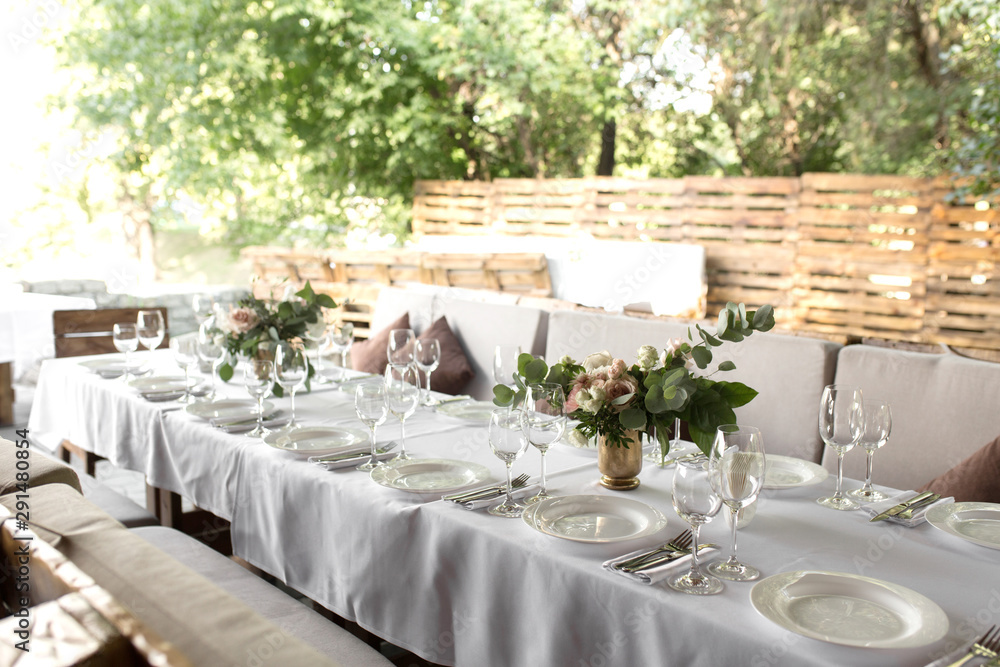 Wedding table setting decorated with fresh flowers in a brass vase. Wedding floristry. Banquet table for guests outdoors with a view of green nature. Bouquet with roses, eustoma and eucalyptus leaves