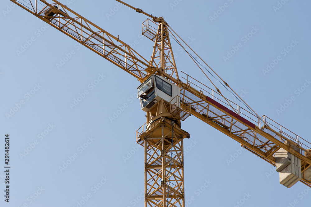 A yellow high-rise building crane against a blue sky builds multi-storey apartment buildings using modern technologies of metal, concrete and brick according to an architectural project A yellow high-rise building crane against a blue sky builds multi-storey apartment buildings using modern technologies of metal, concrete and brick according to an architectural project