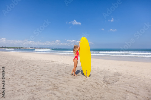 Side view of standing sexy young woman with long hair in red swimsuit with yellow surfboard alone on the beach with waves on Bali island, Indonesia. Active lifestyle. Holiday leisure. Blue sky.