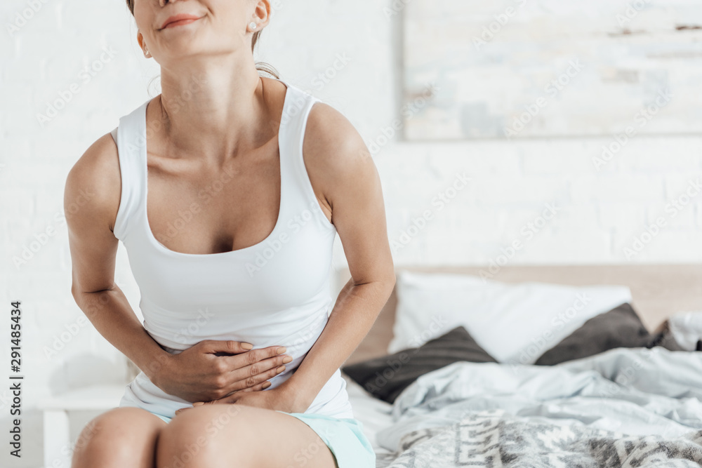partial view of young woman sitting on bed and touching belly partial view of young woman sitting on bed and touching belly