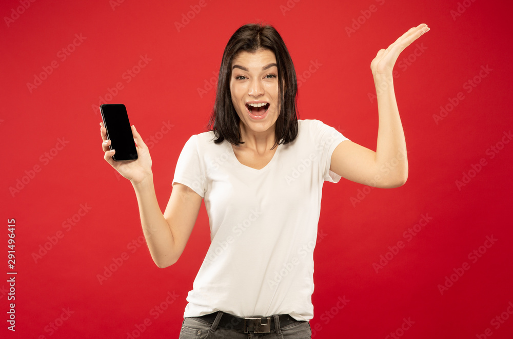 Caucasian young woman's half-length portrait on red studio background. Beautiful female model in white shirt. Concept of human emotions, facial expression, sales. Showing with phone, looks happy. Caucasian young woman's half-length portrait on red studio background. Beautiful female model in white shirt. Concept of human emotions, facial expression, sales. Showing with phone, looks happy.