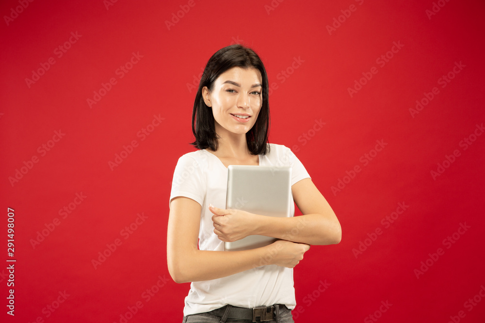 Caucasian young woman's half-length portrait on red studio background. Beautiful female model in white shirt. Concept of human emotions, facial expression. Holding tablet like student or office worker Caucasian young woman's half-length portrait on red studio background. Beautiful female model in white shirt. Concept of human emotions, facial expression. Holding tablet like student or office worker