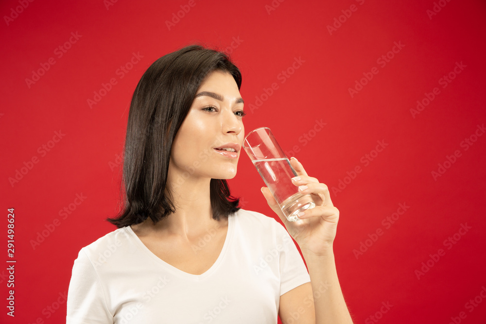 Caucasian young woman's half-length portrait on red studio background. Beautiful female model in shirt. Concept of human emotions, facial expression. Enjoys drinking pure water. Healthy lifestyle. Caucasian young woman's half-length portrait on red studio background. Beautiful female model in shirt. Concept of human emotions, facial expression. Enjoys drinking pure water. Healthy lifestyle.