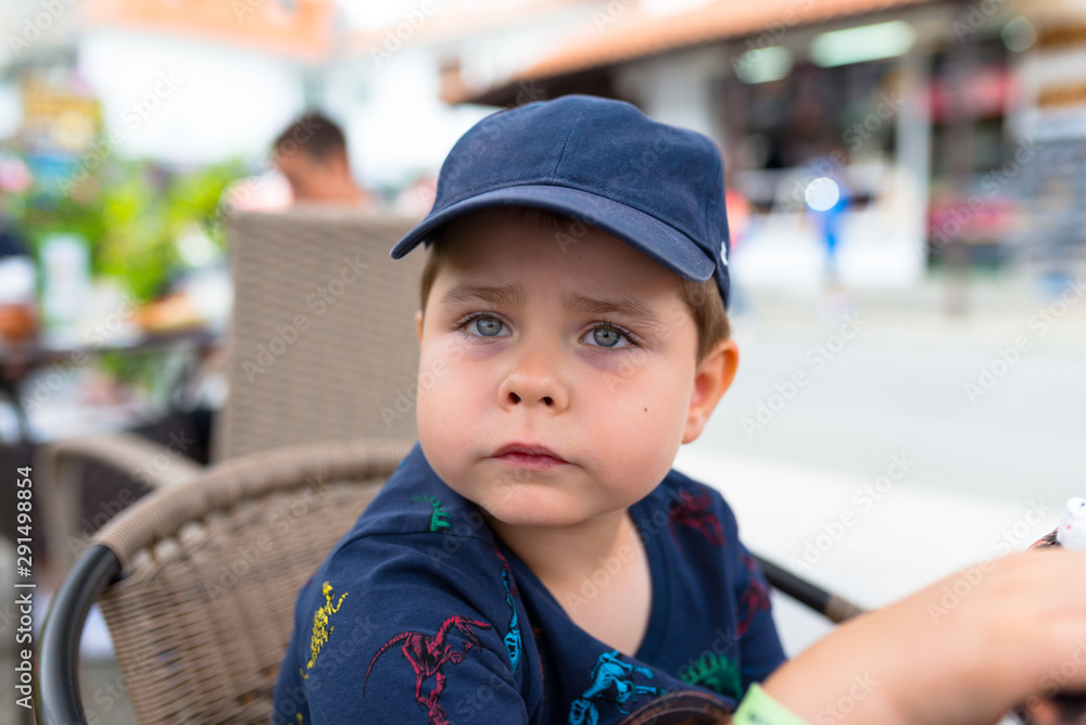 Five-year cute boy in a navy blue baseball cap sitting on a wicker chair outside in a restaurant. Five-year cute boy in a navy blue baseball cap sitting on a wicker chair outside in a restaurant.