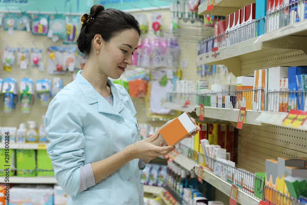 A woman pharmacist in a lab coat with a drug in his hands in a pharmacy near the window with a variety of pharmaceutical products.
