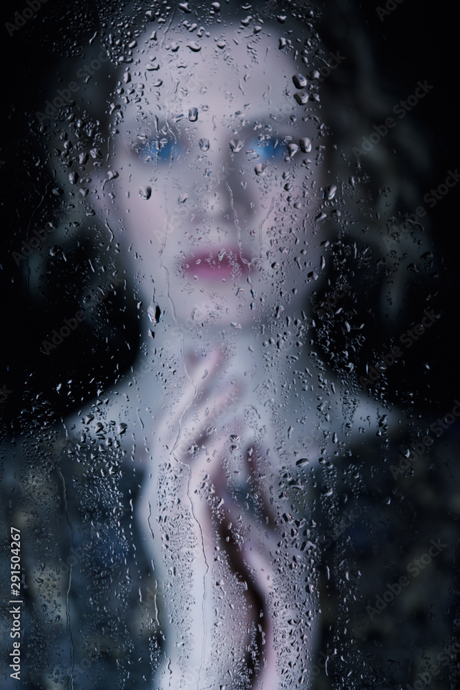 Sad young woman behind the window glass with raindrops
