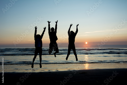 Silhouettes of three teanagers jumping out joyfully on the beach with setting sun at the bakcground