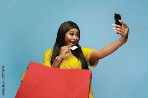 Young african-american woman shopping with colorful packs on blue background. Attractive female model. Finance, black friday, sales, concept. Copyspace. Making selfie or vlog with payment card.