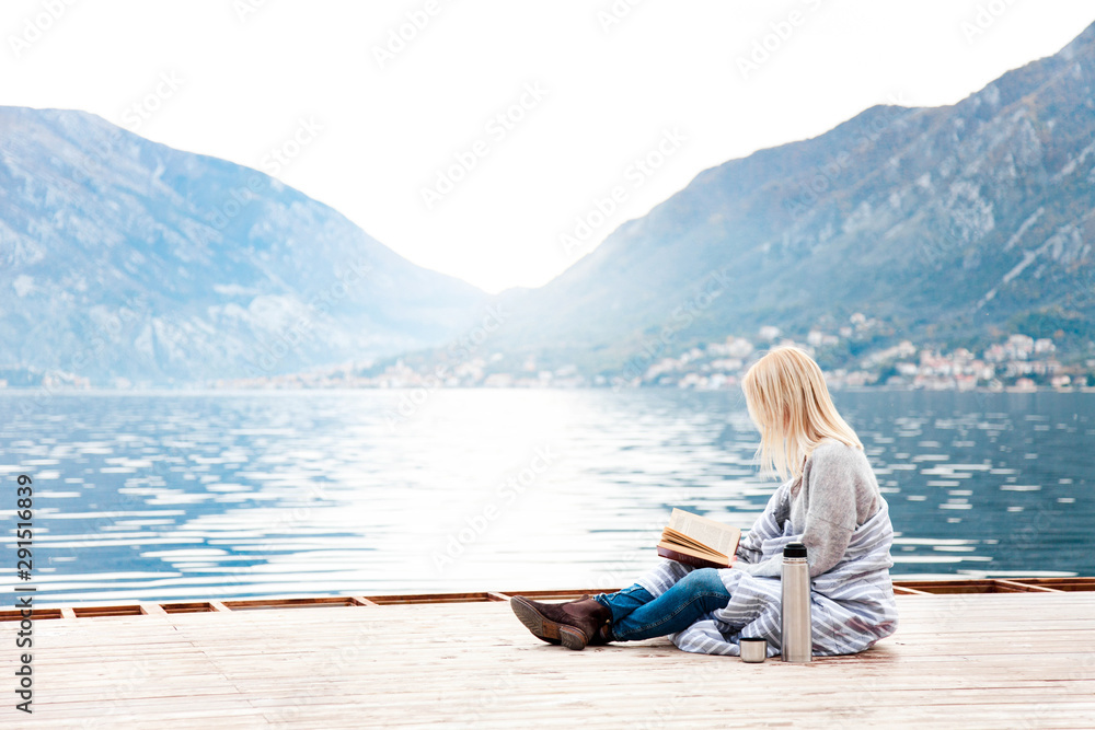 Fototapeta premium Woman is reading book on wooden pier by sea, mountains, beach. Cozy winter picnic with coffee, hot beverages or tea in thermos, warm plaid. Girl is enjoying life, relaxation, wellbeing.