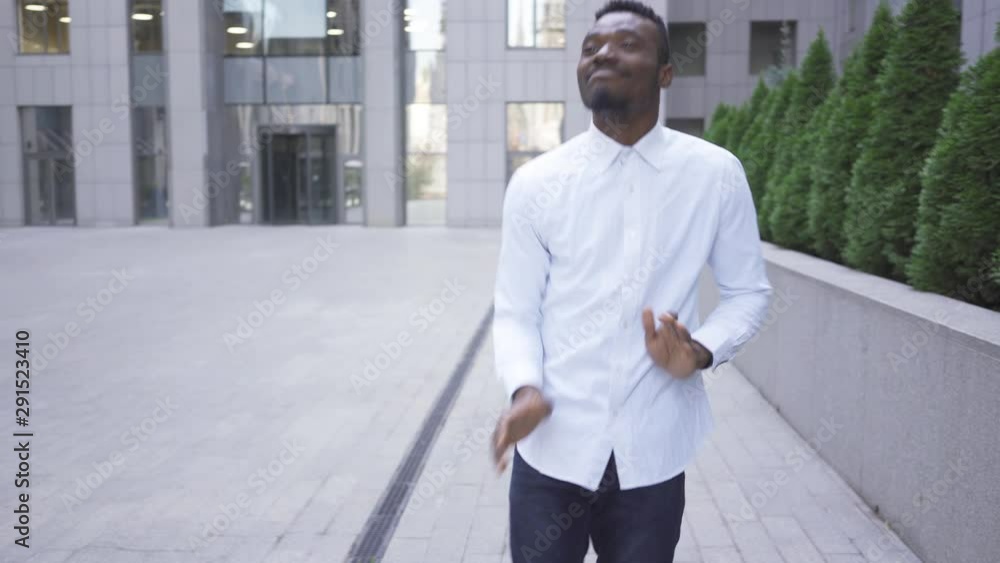 Satisfied African American man in white shirt dancing in front of modern business center on the city street. Male businessman or manager got great news. Satisfied African American man in white shirt dancing in front of modern business center on the city street. Male businessman or manager got great news.