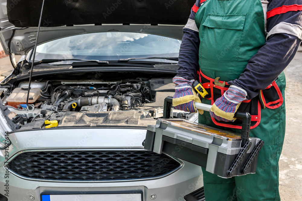 Mechanic in uniform posing with tool box near car engine Mechanic in uniform posing with tool box near car engine