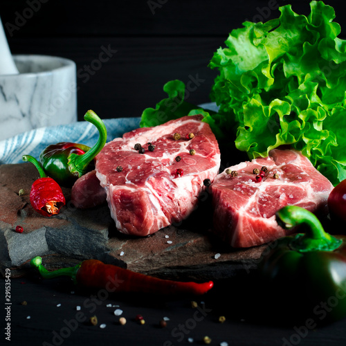 Two juicy steaks lie on a wild stone next to red and green chili peppers and a fresh green salad on a black wooden table