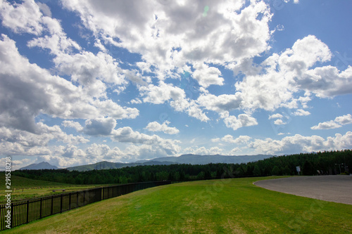 Landscape of green grass with fence, big white clouds, blue sky, and mountain lines from far away in the back - Hokkaido, Japan