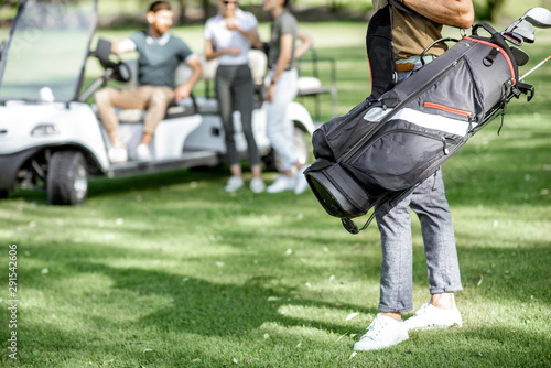 Man standing with golf bag full of putters on the playing course with friends on the background, Close-up view focused on the bag