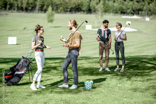Young and elegant friends standing together with golf equipment, having fun during a golf play on the beautiful course on a sunny day