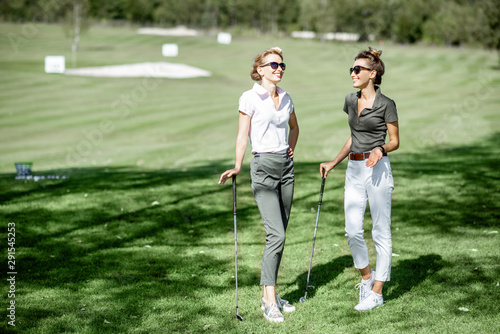 Two female best friends walking with golf equipment on beautiful playing course, talking and having fun during a game on a sunny day