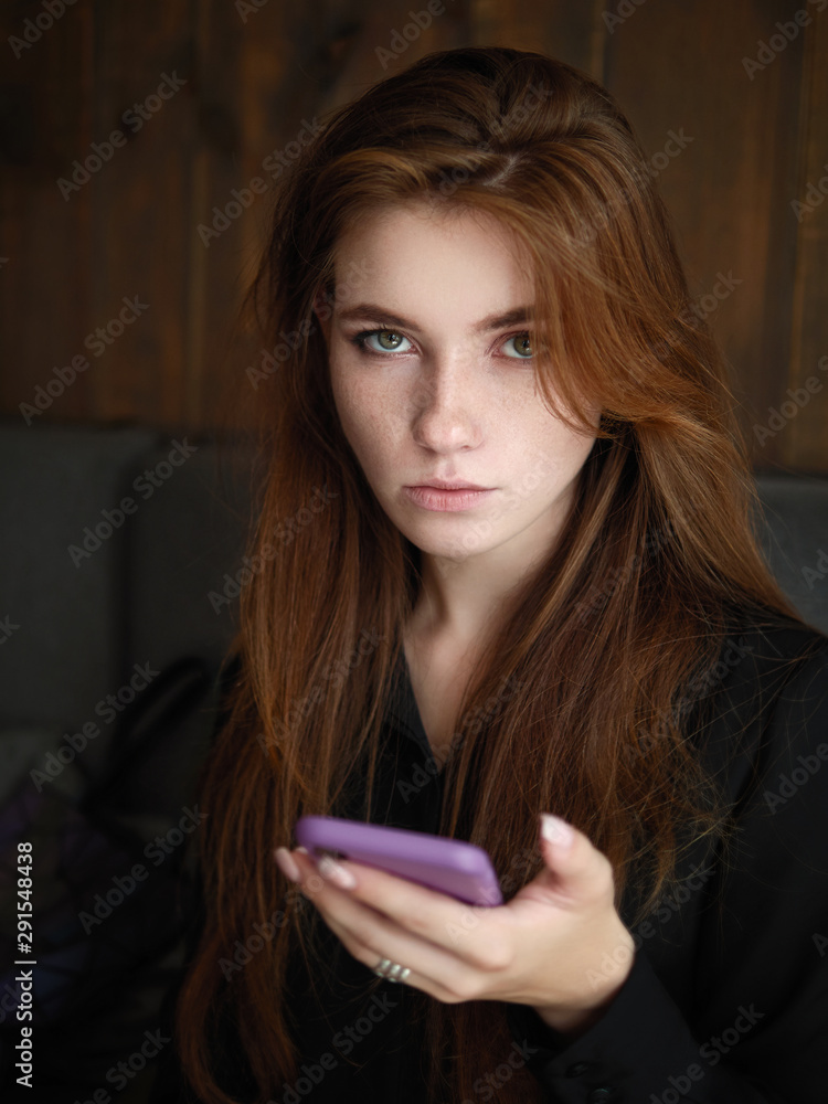 Closeup image of a beautiful serious caucasian red haired woman holding smartphone with adorable smile and feeling good in trendy city cafe Closeup image of a beautiful serious caucasian red haired woman holding smartphone with adorable smile and feeling good in trendy city cafe